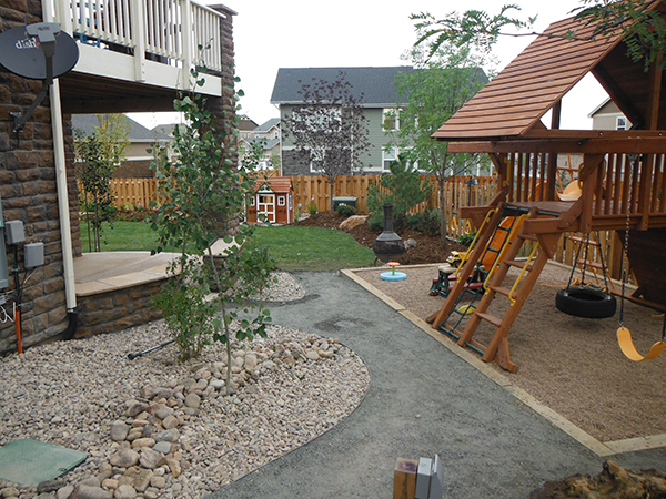 DSCF0276 Backyard with a play structure, swings, slide, and climbing wall on gravel; a small tree and rocks line a curved path; a fenced yard with grass, plants, and playhouse in the distance.