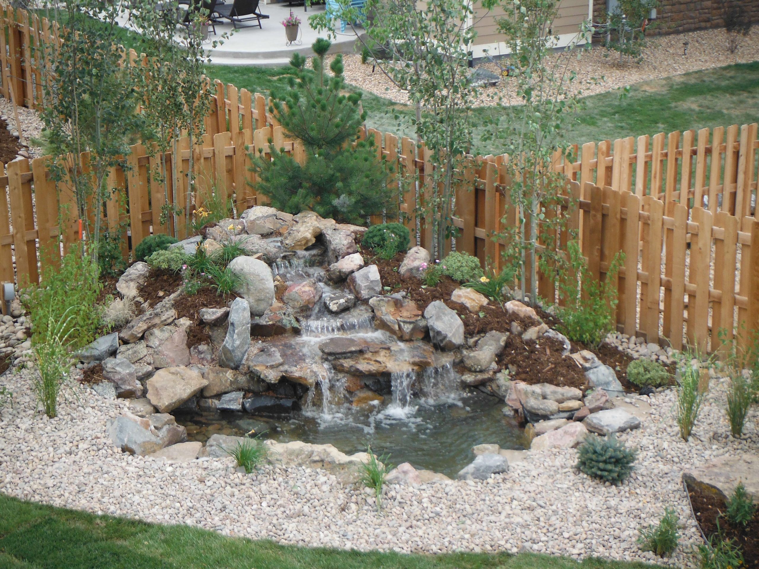 A backyard water feature with cascading small waterfalls flows over rocks into a pond, surrounded by mulch, plants, and a wooden fence. Green grass and other landscaping are visible in the background.