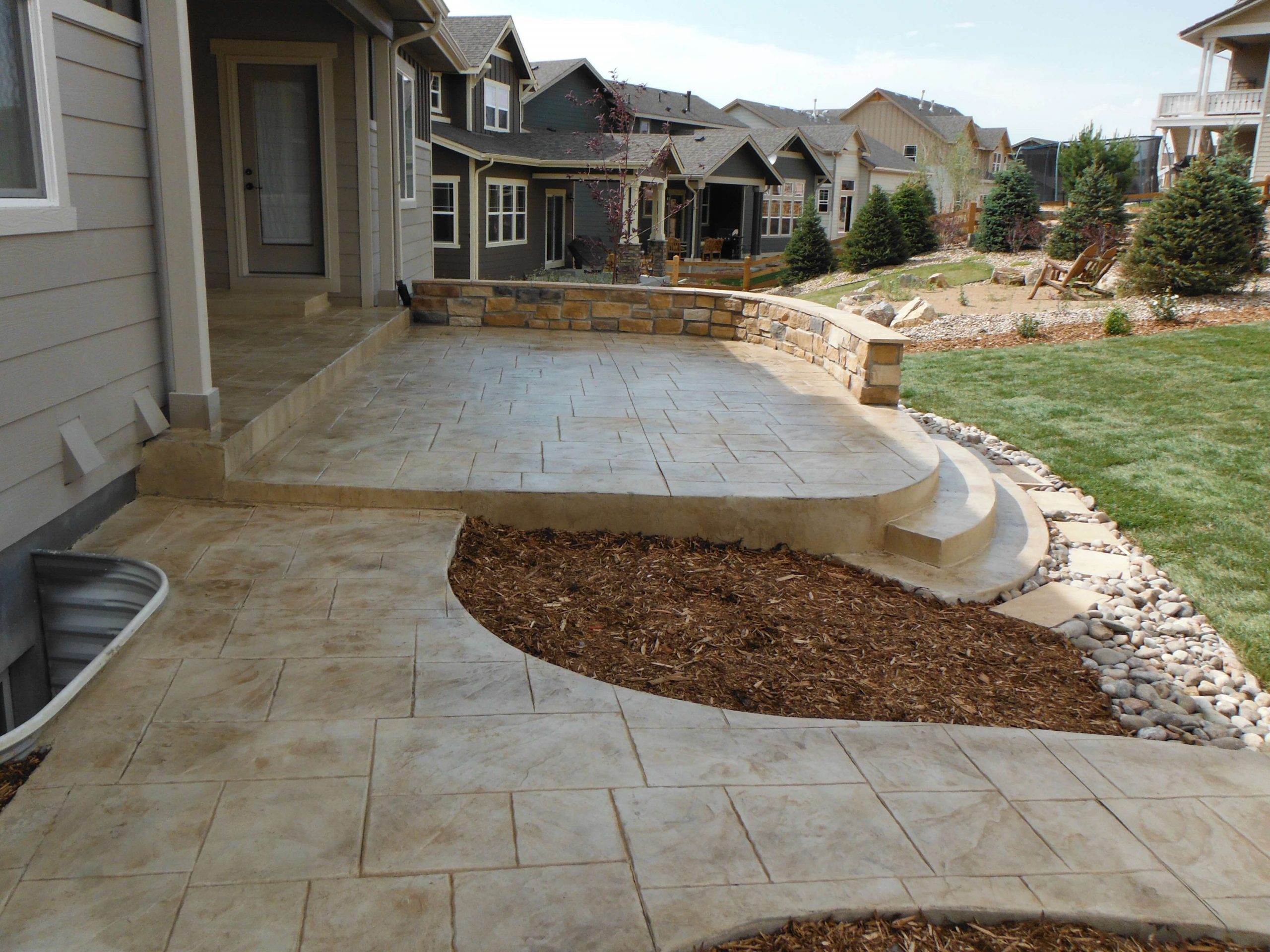 A stamped concrete patio with curved steps, a stone retaining wall, and adjacent mulched and grassy landscaping, situated in a suburban backyard with neighboring houses visible.