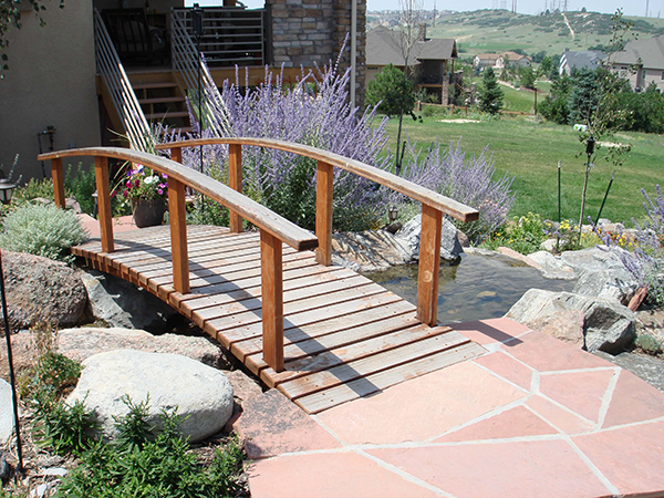 landscape-pics-010 A small wooden footbridge crosses over a narrow pond in a landscaped garden, surrounded by rocks, flowering plants, and a stone pathway, with a grassy area and houses in the background.