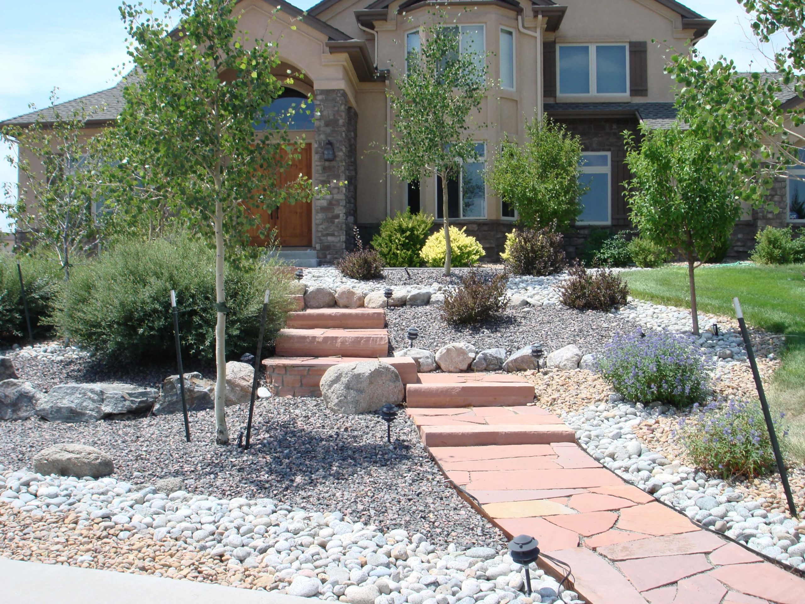 A suburban house with a front yard featuring drought-tolerant landscaping, stone paths, rocks, and small trees and shrubs, with a walkway leading up to the front door.