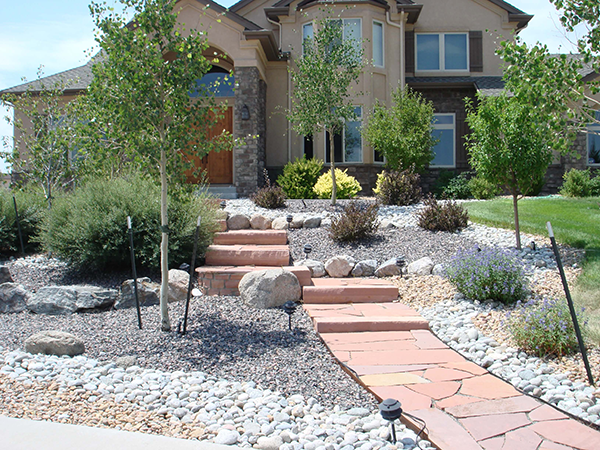 landscape-pics-017 A landscaped front yard with a stone path, terraced steps, rocks, mulch, shrubs, and small trees leading to the entrance of a tan house with large windows.