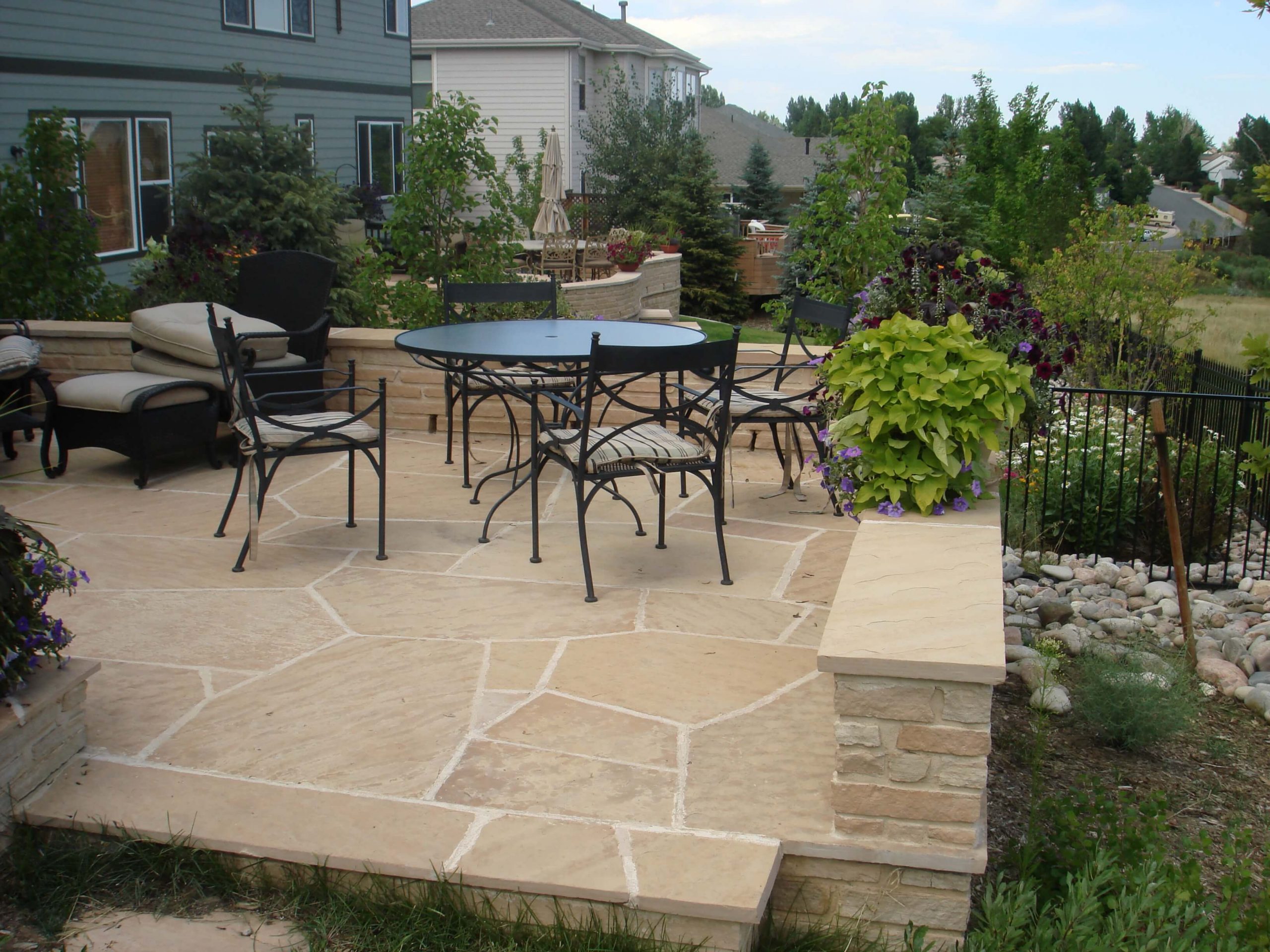 A patio with beige stone flooring, black metal chairs and a round table, surrounded by greenery and potted plants, in a suburban backyard next to houses and a landscaped garden.