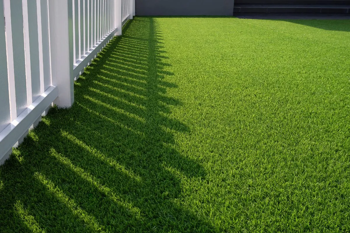 A close-up of a vibrant green artificial grass lawn next to a white fence, with the fence casting long, distinct shadows across the grass in bright sunlight.
