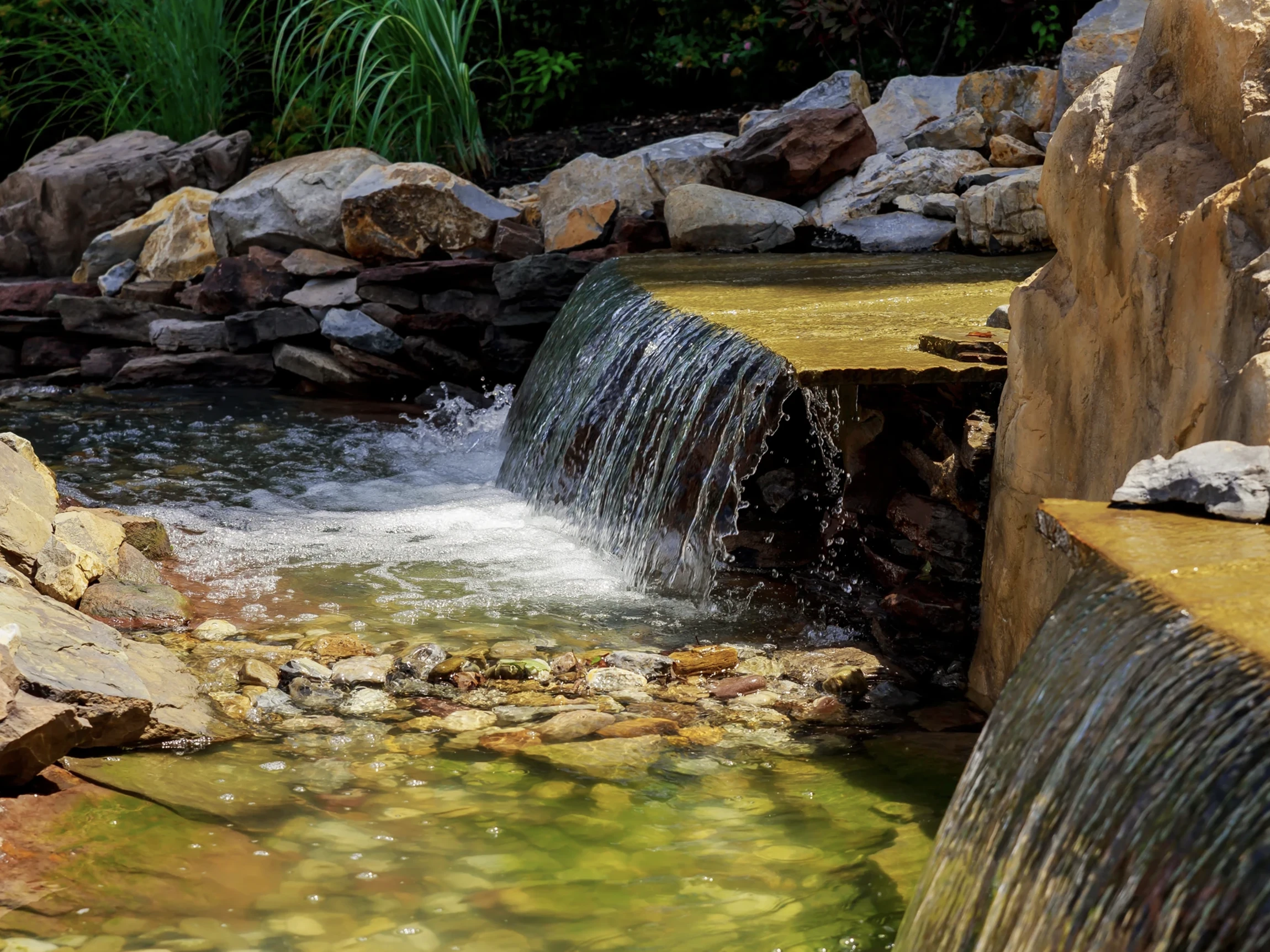 Small waterfall flows over flat rocks into a clear, shallow pond surrounded by stones and green plants, creating a serene garden scene.