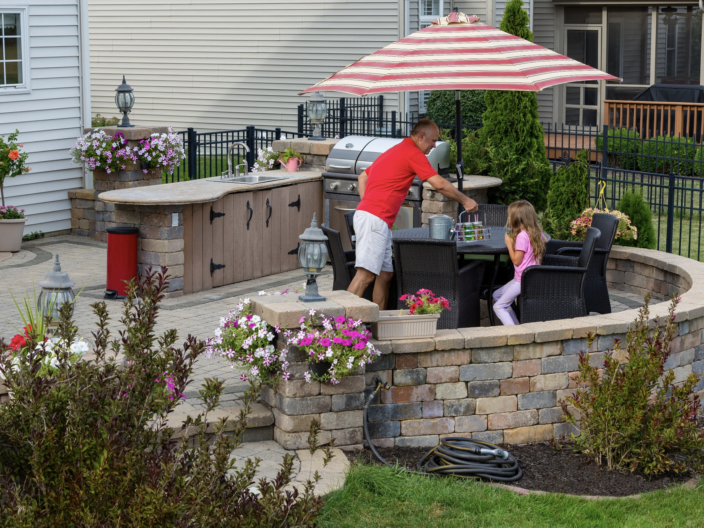 A man in a red shirt serves food to a young girl at a patio table under a striped umbrella. The patio has a built-in grill, potted flowers, and is surrounded by a stone wall and landscaping.