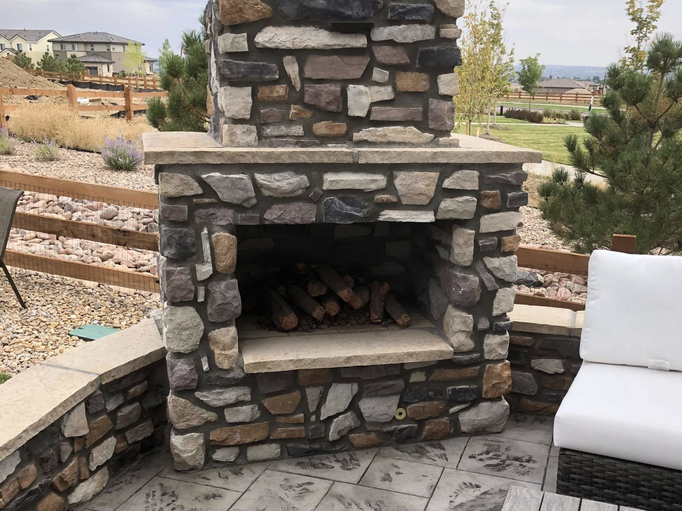 A stone outdoor fireplace with stacked firewood sits on a patio. A white cushioned chair is partially visible nearby, and the area is surrounded by landscaping, a wooden fence, and green lawns.