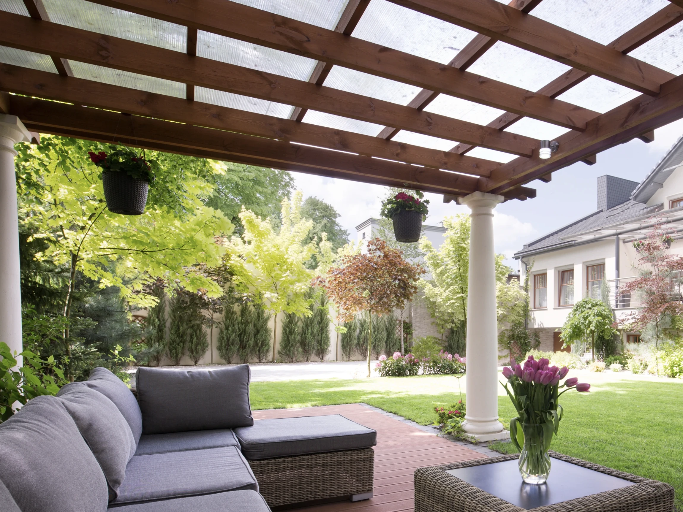 A cozy patio with a gray sectional sofa and coffee table sits under a pergola with hanging plants, overlooking a lush green garden and a modern house in the background.