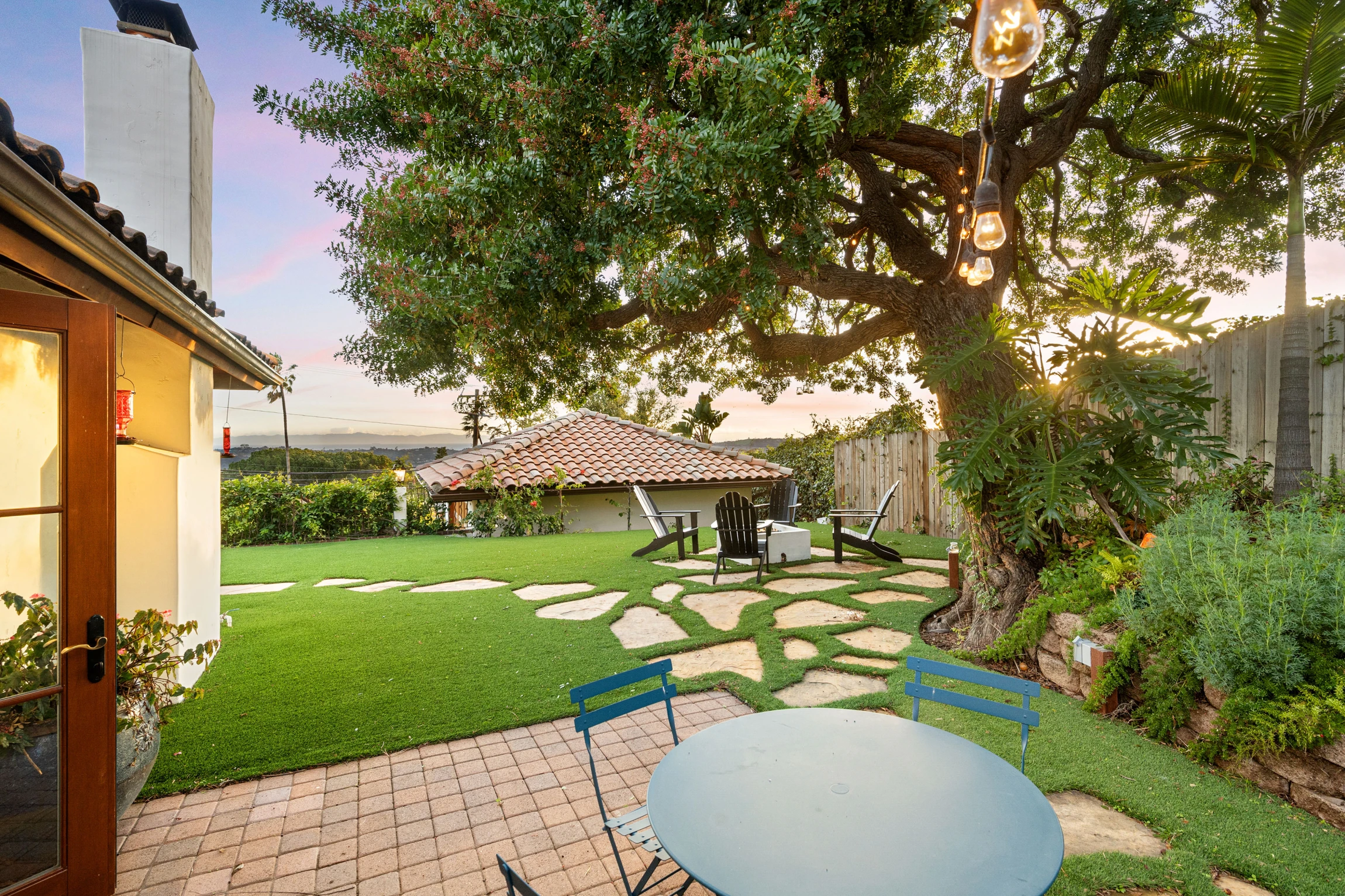 A sunny backyard with a round blue table and chairs on a stone patio, string lights hanging from a large leafy tree, stone path, green lawn, and Adirondack chairs around a fire pit near a tiled-roof structure.